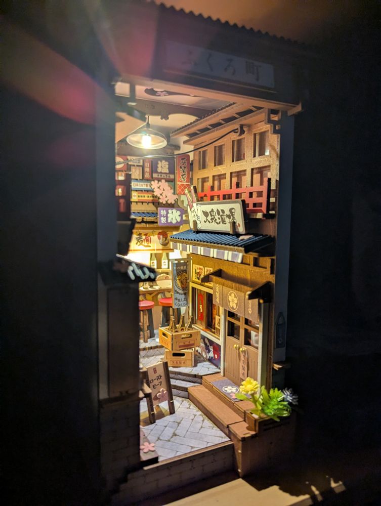 Book nook diorama of a Japanese alley lined with shops and food stalls. The diorama is lit up by various lights located in the diorama.
