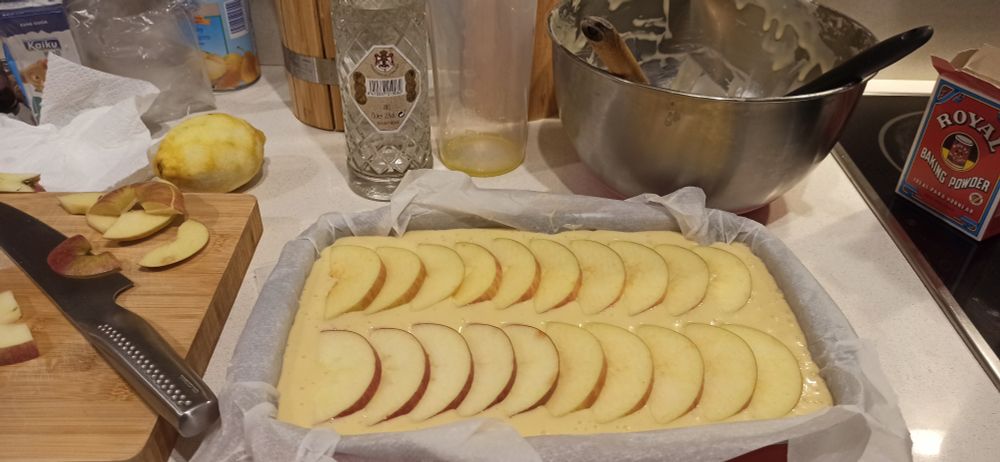 Cóc d'oli amb poma, a punt per entrar al forn. Al voltant s'hi veuen restes de poma, una ampolla d'Anís del Mono i utensilis amb restes dels ingredients.