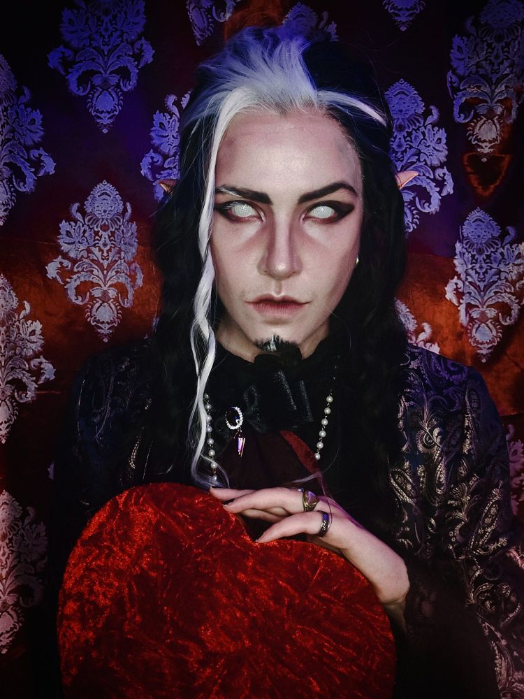 A photo portrait of a vampire with whiteout contacts. White and black hair and pointed ears. They are holding a plush red heart box 