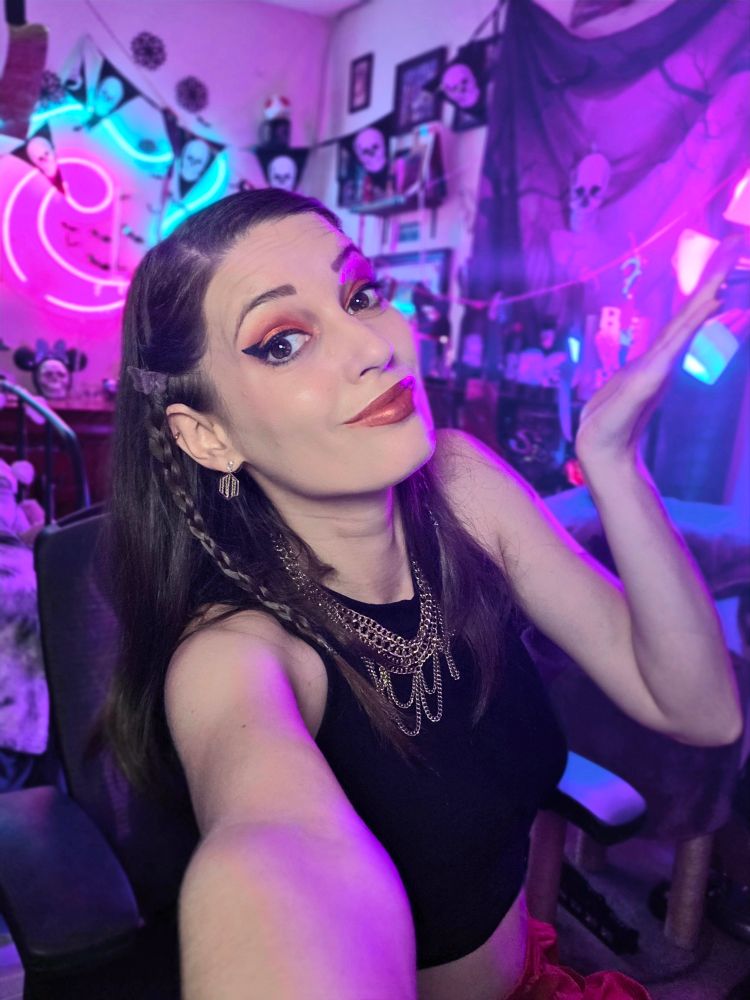 Char shrugging with a smirk in her stream room. Its is decorated with skull flags and a creepy backdrop for Halloween.
