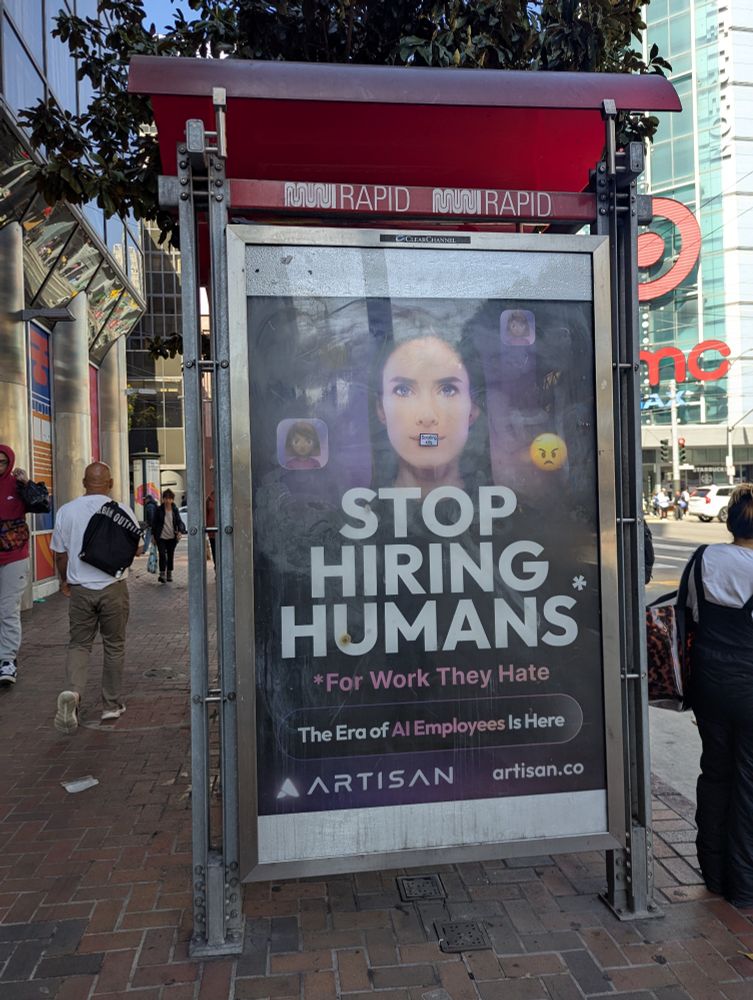 An ad on the side of a bus stop in San Francisco that says "STOP HIRING HUMANS* 
*for work they hate. The Era of AI Employees is here"