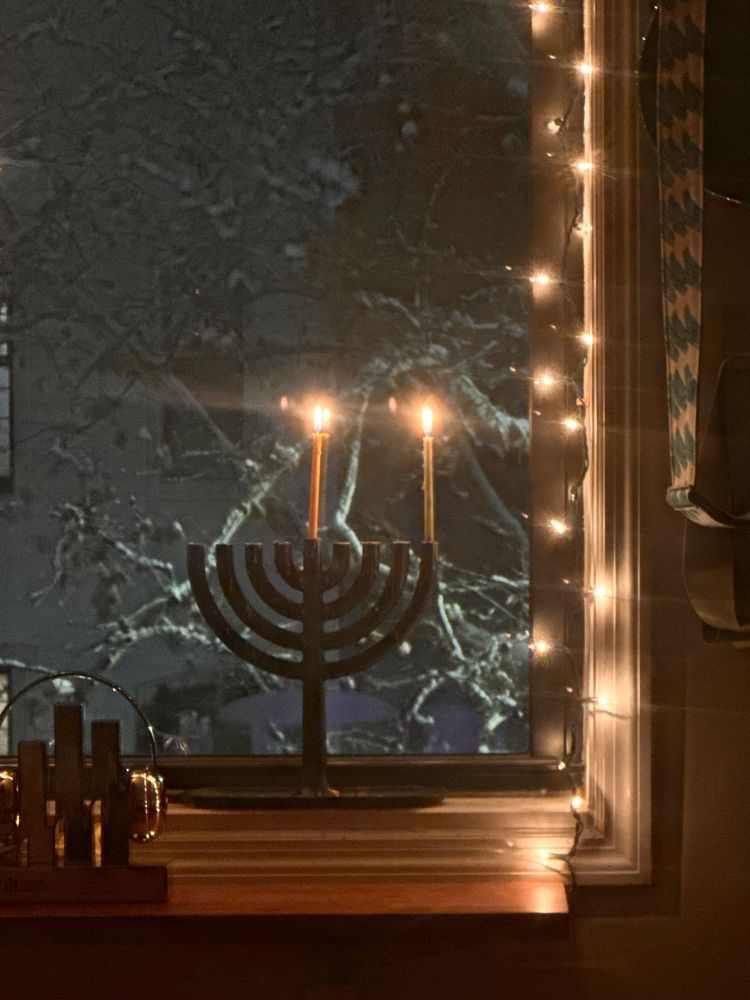 Picture of a black menorah with two candles burningin a window with branches across the street, framed by Christmas lights
