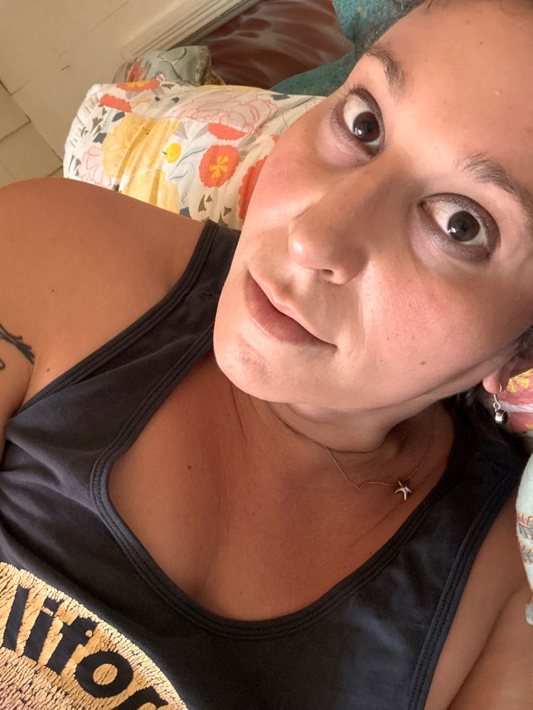 Trans girl, sleepy face and big brown eyes stares at the camera, she’s got a little remnant of last night‘s makeup on and some visible cleavage and is wearing a black tank top