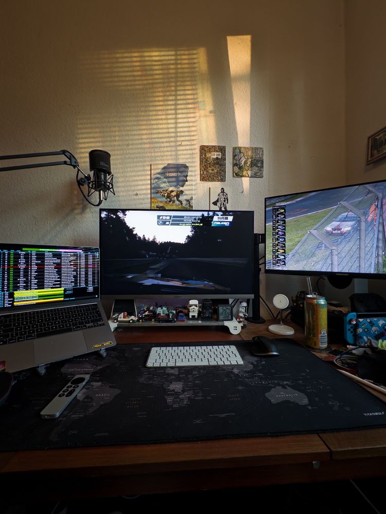 Das Foto zeigt einen aufgeräumten Schreibtisch mit einem modernen Setup, bestehend aus drei Bildschirmen. Auf der linken Seite befindet sich ein aufgeklapptes MacBook mit einem externen Monitor in der Mitte und einem weiteren Bildschirm rechts. Die Monitore zeigen Live-Übertragungen eines Autorennens und Rennergebnisse. Vor den Monitoren steht eine weiße kabellose Tastatur sowie eine Maus auf einem großen Mauspad mit einer Weltkarten-Grafik. Auf dem Schreibtisch liegen außerdem eine Fernbedienung und eine Getränkedose. Im Hintergrund ist ein Mikrofon an einem Schwenkarm befestigt. Die Wand zieren einige Poster und ein Schattenmuster von einem Fenster mit Jalousien, das für ein warmes Lichtspiel sorgt. Unter dem mittleren Monitor stehen kleine Figuren und Fahrzeuge, vermutlich aus Lego.
