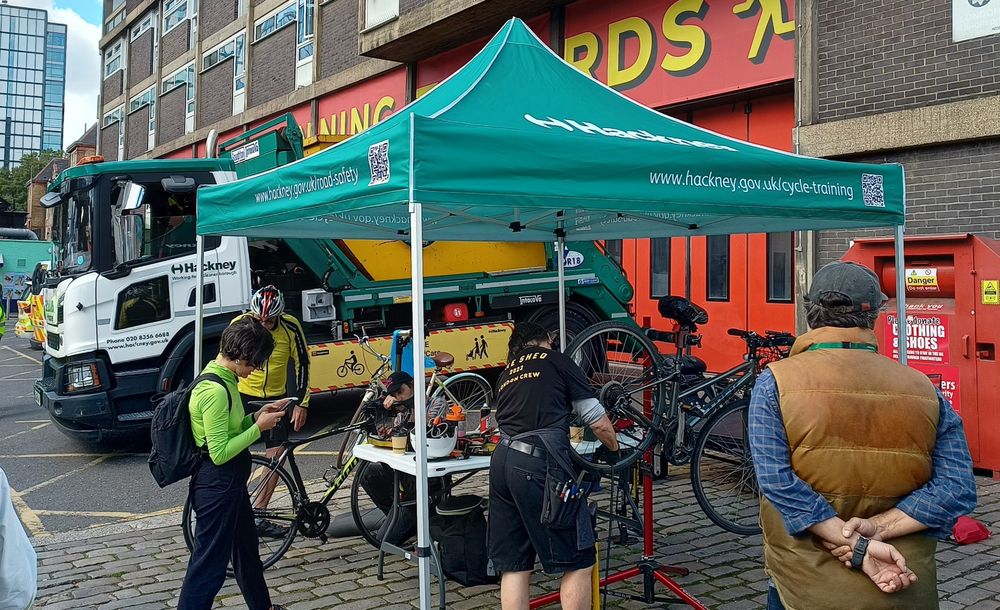 A group of people standing in front of a green gazebo, while a mechanic checks a bike