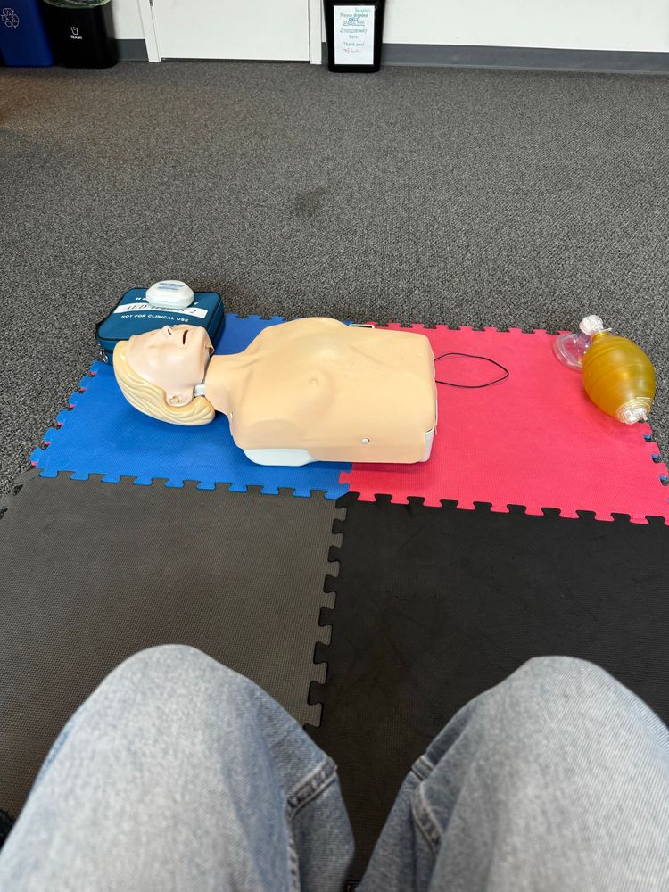 A cpr manikin laying on the floor with a training aed and ambubag nearby