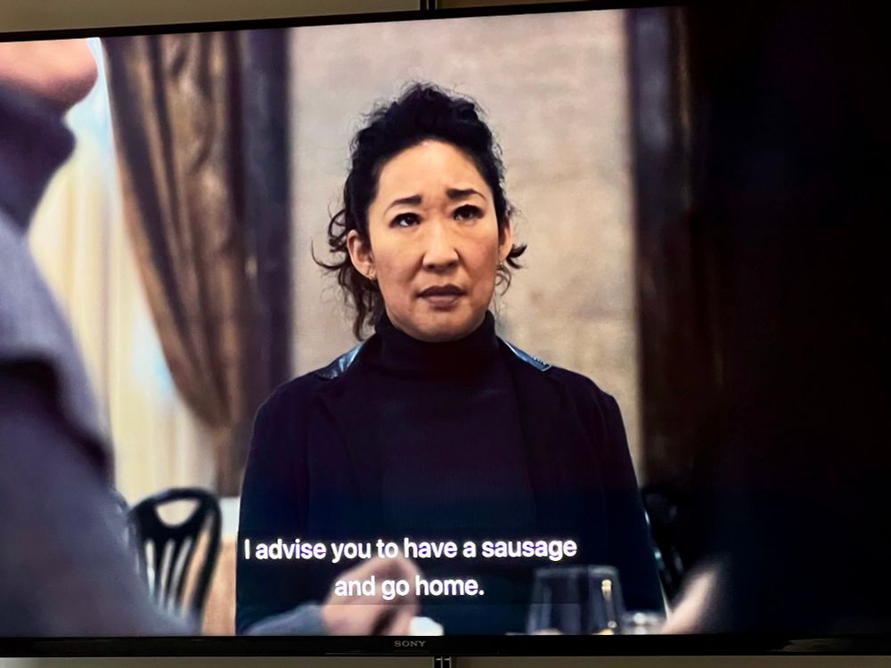 An out-of-context still image from the TV programme “Killing Eve”. A woman is looking off centre camera as someone opposite is speaking to her. The subtitles read: “I advise you to have a sausage and go home.”