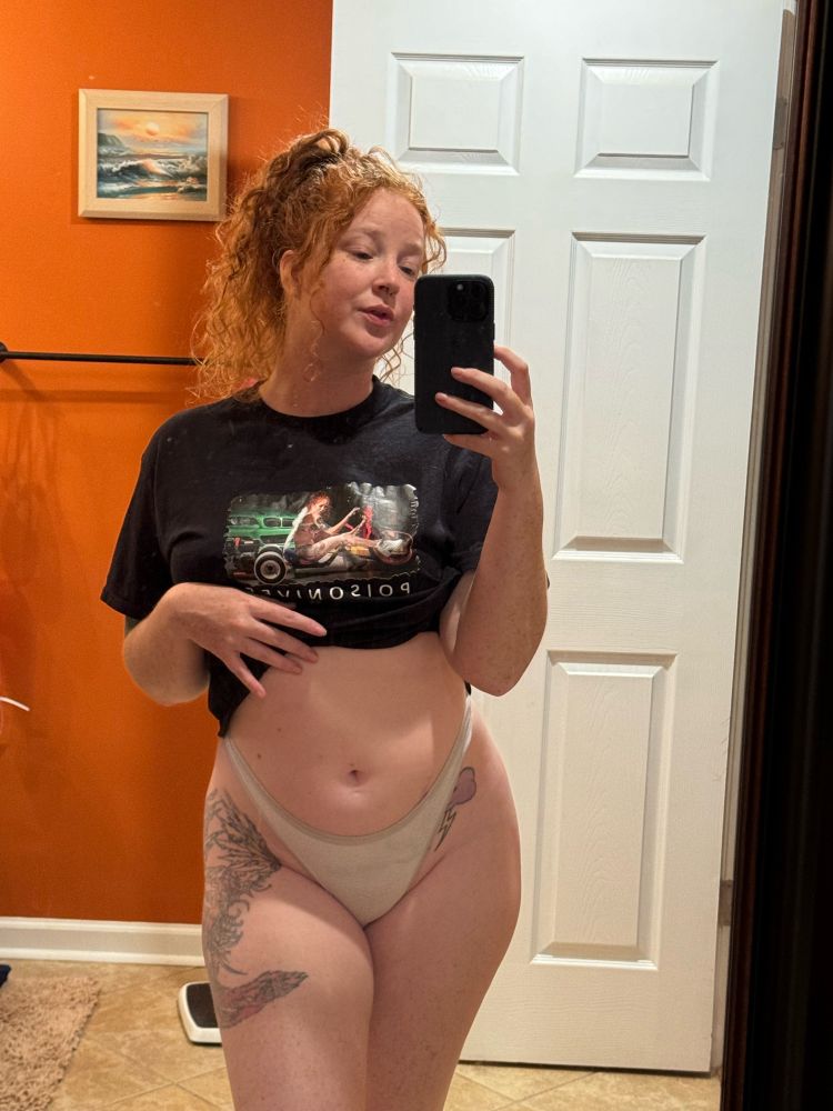 Woman with curly red hair up in a ponytail, taking a picture of her reflection from the knees up, wearing pulling her shirt up to show off her stomach and white cotton thong panties.