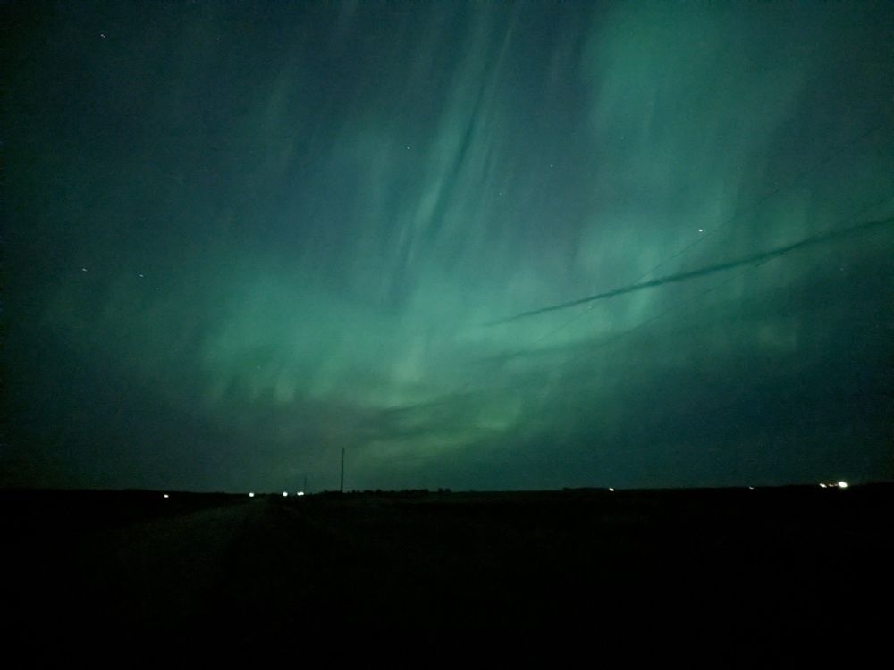 Greenish northern lights above a flat,black horizon with an airplane's contrail cutting through it