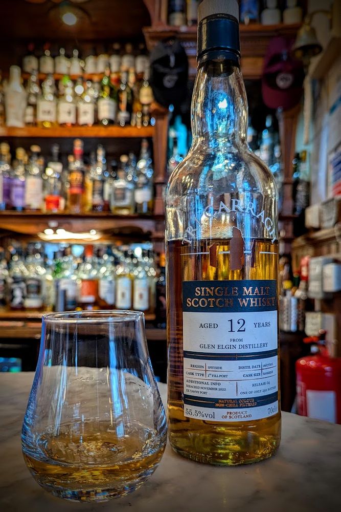 A bottle of Glen Elgin whisky on a bar top beside a glass with a measure. Shelves of whisky bottles in the background.