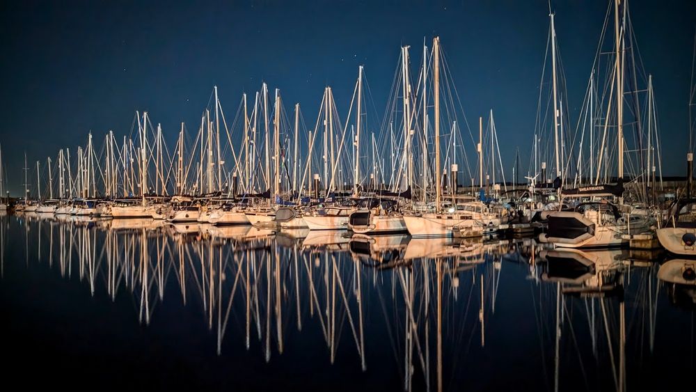 A shot of yachts in a marina on a clear night with their masts hulls reflected in still water.