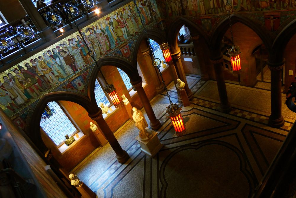 A photograph looking down on the Foyer of the Scottish National Portrait Gallery. A tiled floor with arches around each side supporting an upper gallery. A statue of Robert Burns in the Foyer.
