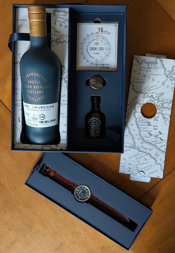 An open gift box on a wooden table containing whisky and a watch.