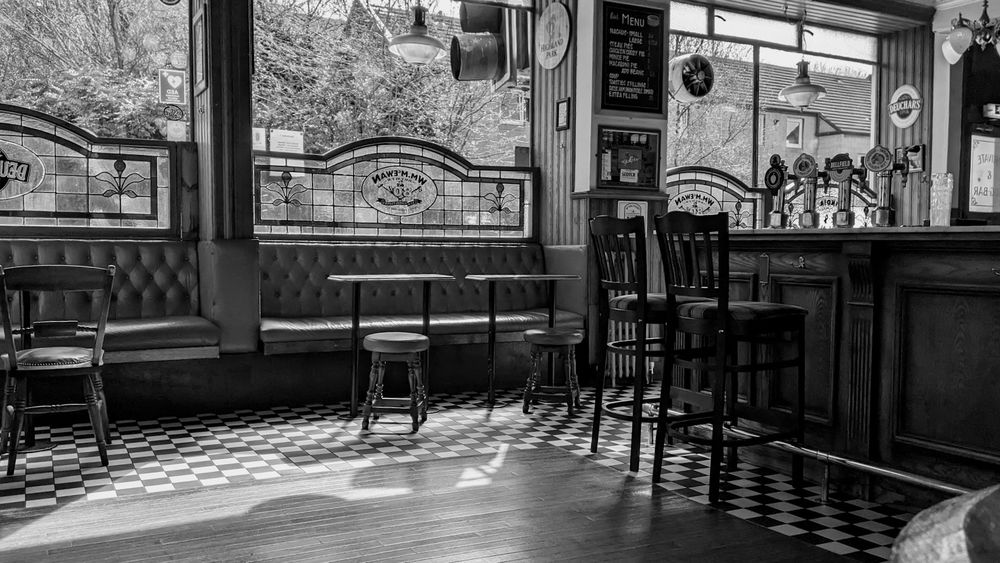 A black and white photo of the interior of a pub showing a bar, tables and chairs with sun shining through windows.