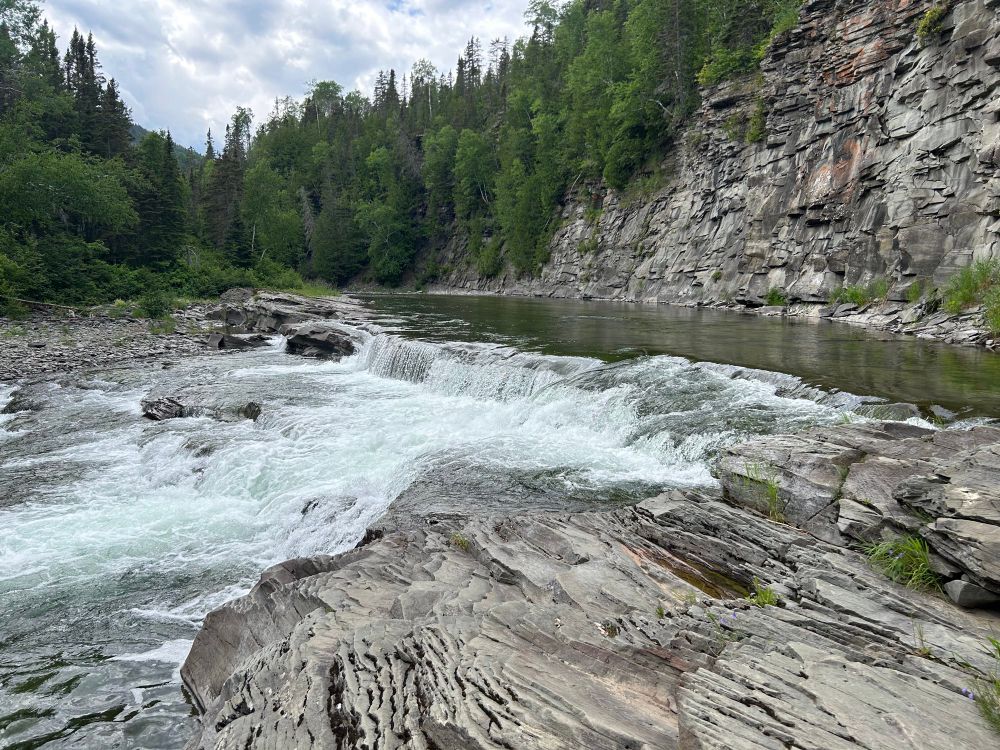 A cascade in the river. On the right a steep cliff has been carved by the river. The rock of the cliff and foreground is relatively thin layers of shale. 