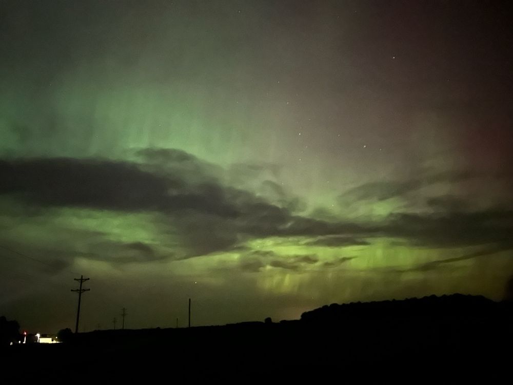 A night sky scape looking north: a farm with lights in the distant horizon. Above a dark narrow cloud runs horizontal across the image, but green and purple streaks radiate above and below it.