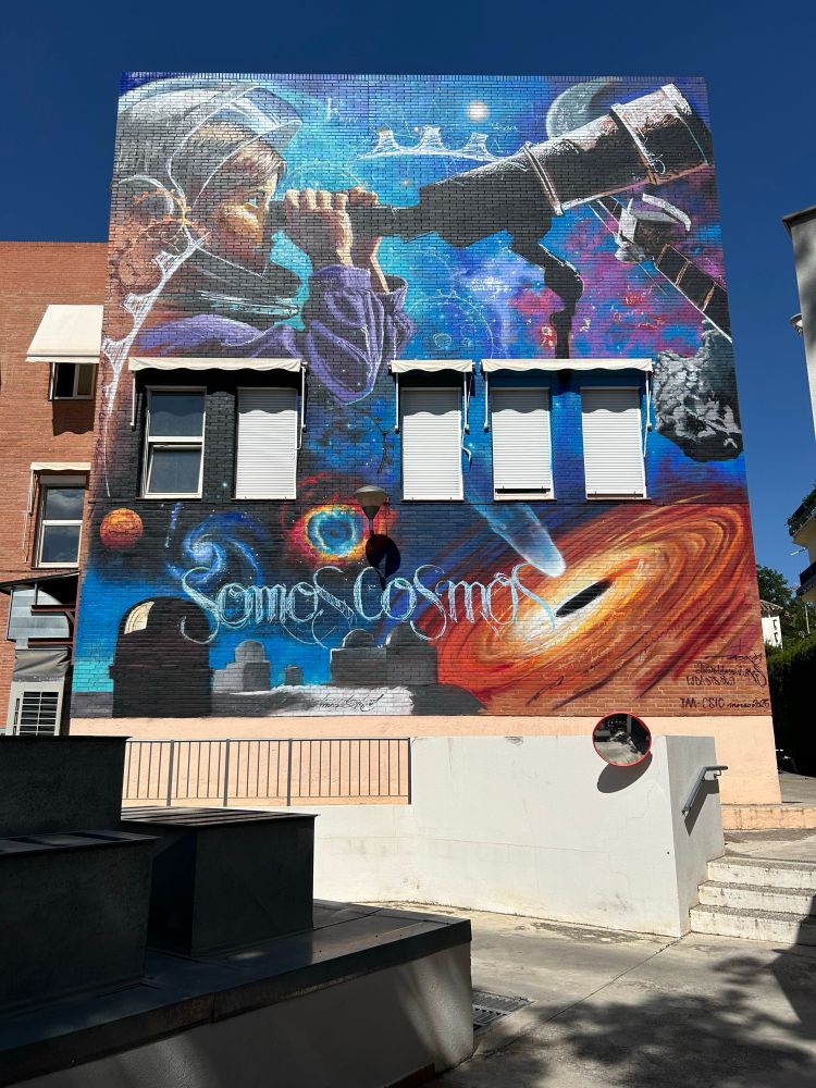 Mural on the south side of a two story building facing south. The mural surrounds five windows with their blinds drawn. Spanning the top of the mural, a girl in an astronaut helmet looks through a telescope. At the bottom of the image and in the background you see astronomical objects like a black hole with accretion disk and jets, planetary nebula, a galaxy and planet. A tribute to the ground and space based telescopes that the IAA contributes instrumentation to are also there. The colors are bright and in the artist’s signature style are the words “somos cosmos” in the lower half. 