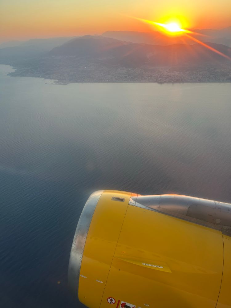 Sunset from an airplane off the south coast of Spain. The fuselage of a plane is seen against a relatively calm sea with the sun setting behind mountains on the coast. 