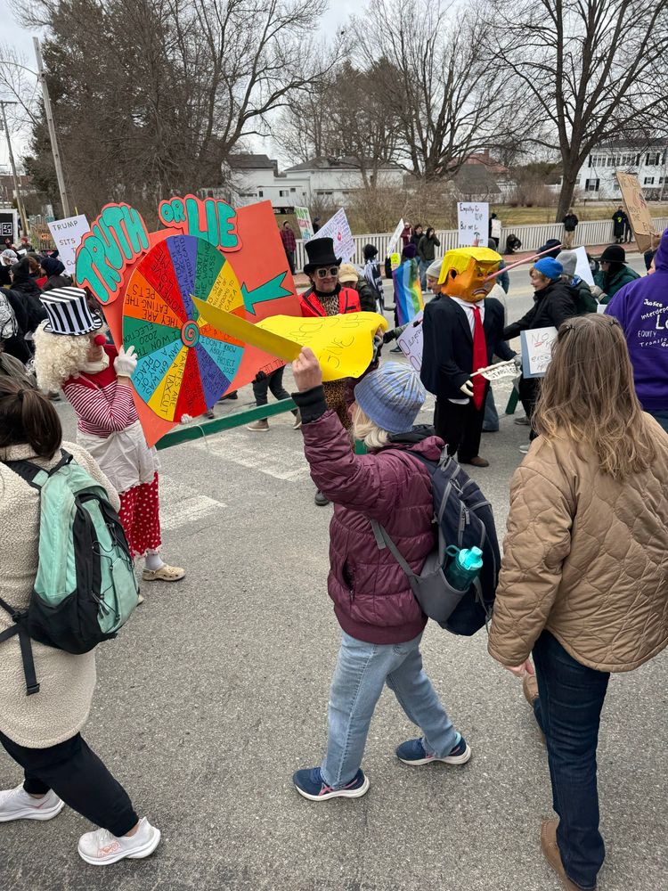 Photo of giant game board with spinning wheel, truth or lies. Next to this is a person in a suit with a papier-mâché head decorated to look like Trump. Where his nose should be is a very long pointed stick; it is Pinocchio like.