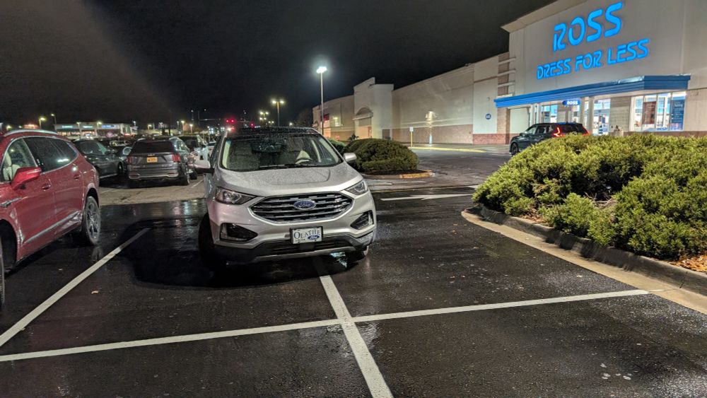 Small white SUV poorly parked, fully blocking two spots in the front row of parking at a rainy strip mall 