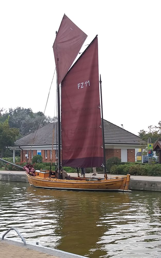 Das Bild zeigt ein Segelboot aus Holz mit dunkelroten Segeln von der Seite. Dahinter ein Gebäude aus rotem Klinker und dunklem Dach. Seitlich des Gebäudes Bäume und Büsche.  Darüber ein grauer Himmel. 