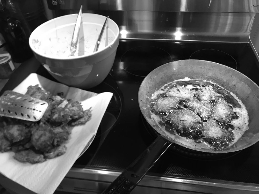 Malanga fritters frying in a pan, with a bowl of batter and finished fritters to the side (in grayscale)