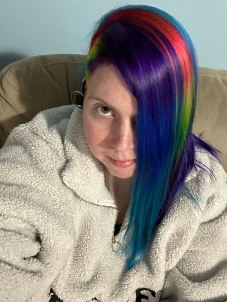 Picture of a white woman with a side shave in rainbow colors. The front of her hair is a dark purple to blue to teal ombré with a rainbow stripe behind it.