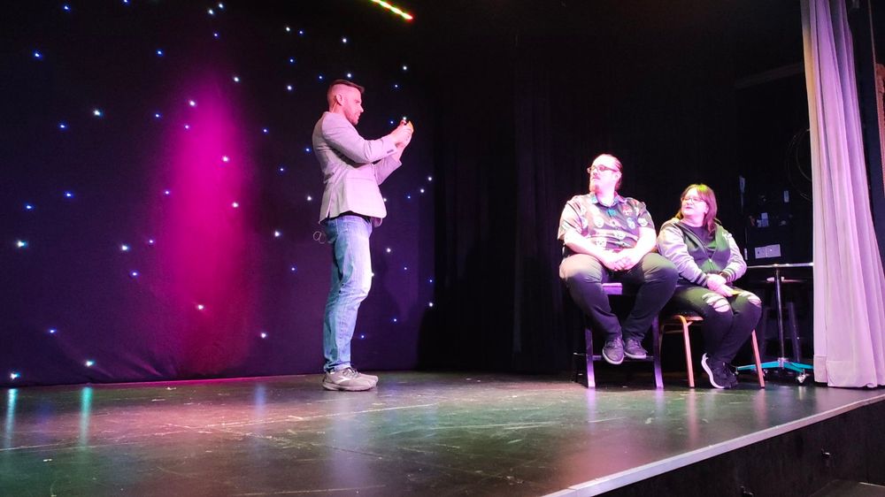 Magician Nigel McGuinness standing on stage with volunteers Joey and Rickee sitting down