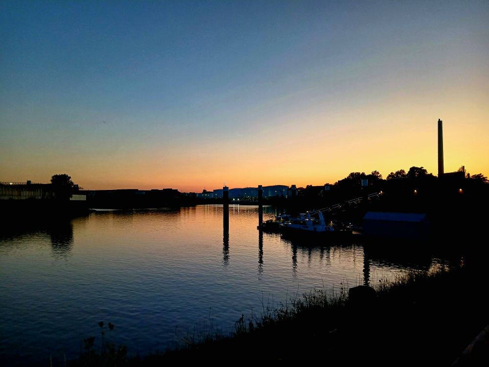 Orange-blauer Abendhimmel über Wasser. Im Hintergrund sind Schornsteine und Industriegebiet zu sehen.