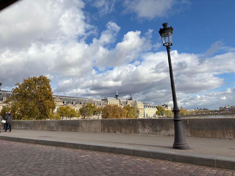 Photo Pont Royal Paris avec le Musée du Louvre en arrière plan. Un lampadaire sur la gauche et le ciel nuageux et bleu 