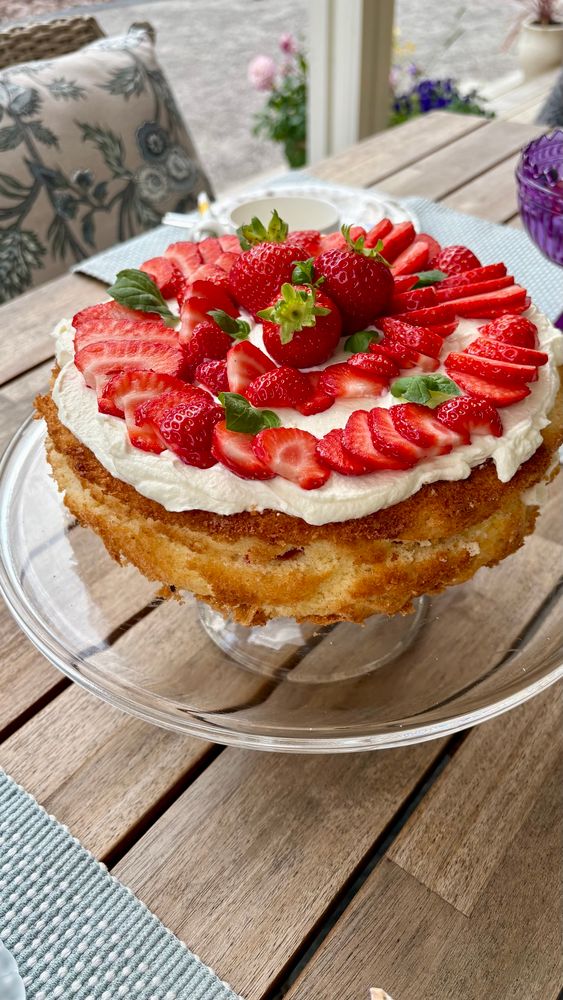 Ricottajuustoa, kermaa, mantelijauhetta ja mansikkaa sisältävä täytekakku on päällystetty mansikkaviipaleilla ja mintunlehdillä. Kakku on jalallisen lasikakkuvadin päällä. Terassilla näkyy myös vaaleanpunaisia ja -sinisiä kesäkukkia.