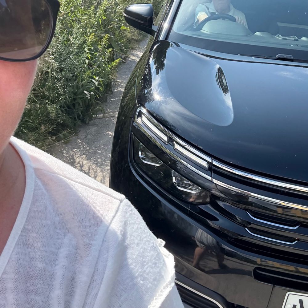 Vanha miesoletettu mustan Citroënin ratissa ajaa jalankulkijoiden kannoilla hiekkatiellä. 