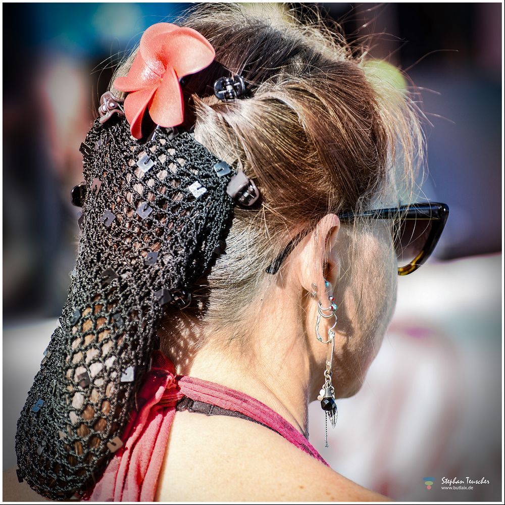 Eine Frau von hinten mit Sonnenbrille, auffälligem Ohrschmuck und Netzhaarnetz, das mit Perlen und einer roten Blume geschmückt ist. Sie trägt ein rotes Oberteil. Die Szene wirkt wie Teil eines Events oder Festivals.