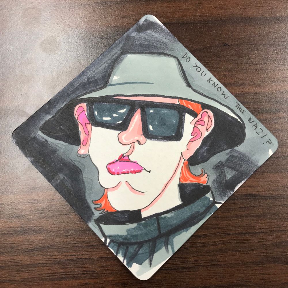 A drawing on a coaster of a bucket hat with a pouty Nazi under it; his glasses hide his dead eyes, but his ginger hair has escaped the confines of his hat.