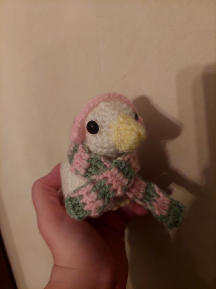 a hand holding a white crochet duck with a green and pink scarf and pink earmuffs 
