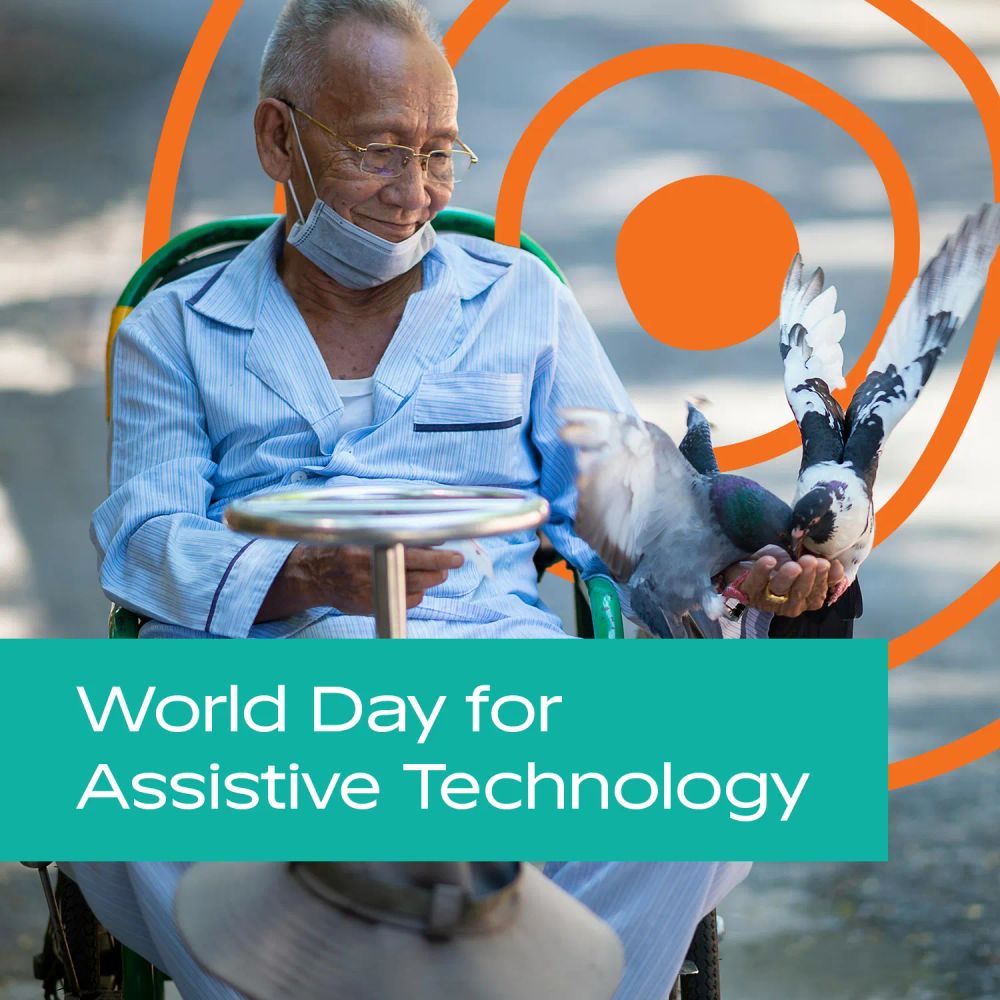 An older man in a wheelchair, feeding pigeons in a park outside whilst smiling. The UN Decade of Healthy Ageing logo is in the background. Text: 'World Day for Assistive Technology'.
