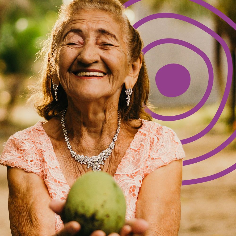An older woman in a rural environment, offering a fruit to the viewer while smiling. The UN Decade of Healthy Ageing logo is in the background.
