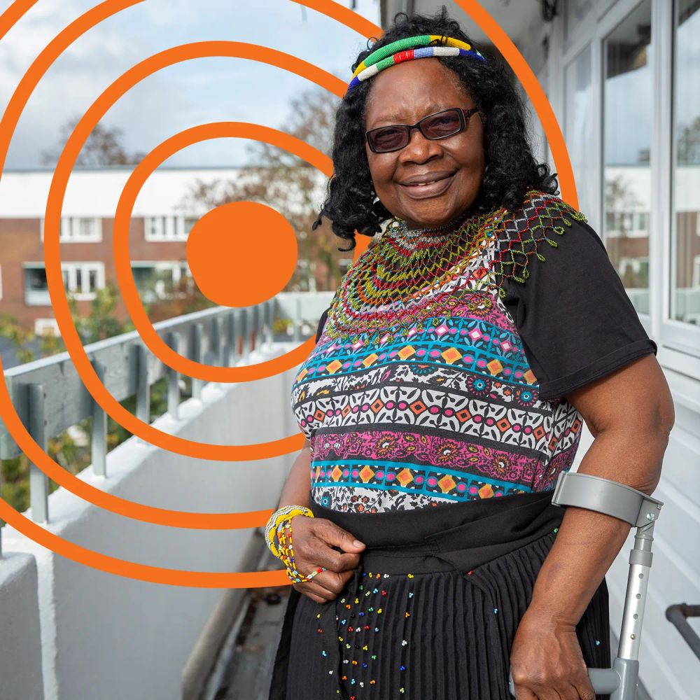 An older woman, wearing colourful clothing, smiling at the camera, on her balcony. She is using an assistive product - a walking stick - to move around. The UN Decade of Healthy Ageing logo is in the background.