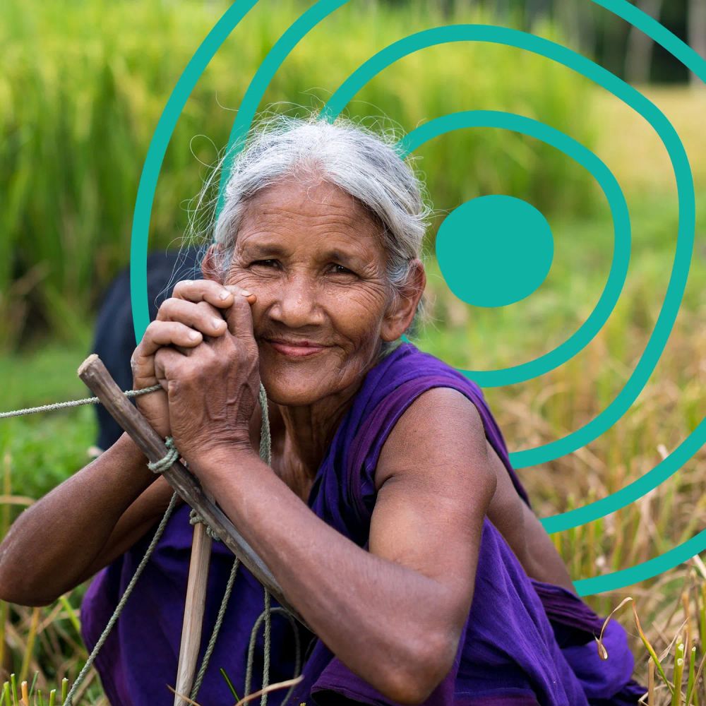 An older woman in a field, squatting down while smiling at the camera with her hands clasped. The UN Decade of Healthy Ageing logo is in the background.