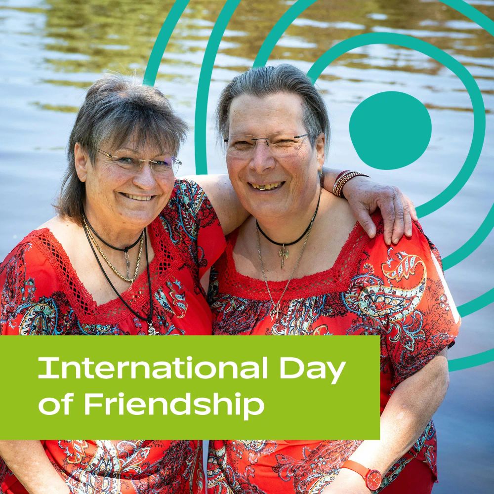 Two older friends wearing similar outfits, on a day out outdoors. Text: 'International Day of Friendship'