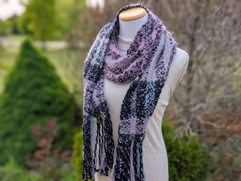 A purple, blue, and black woven scarf draped over a mannequin with shrubs blurred out in the background.
