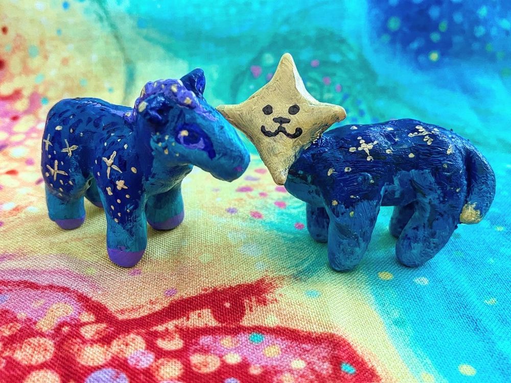 A pair of handmade sculptures (made with polymer clay and painted with acrylic paint) of a starry blue horse and a creature with a star head and a :3 face
