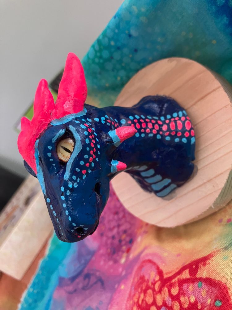 A hand sculpted dragon made of polymer clay, painted with acrylic paint, and mounted on a recycled wood board. Done in a faux taxidermy head mount style.