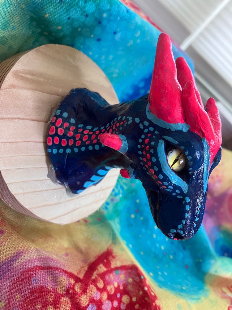 A hand sculpted dragon made of polymer clay, painted with acrylic paint, and mounted on a recycled wood board. Done in a faux taxidermy head mount style. 