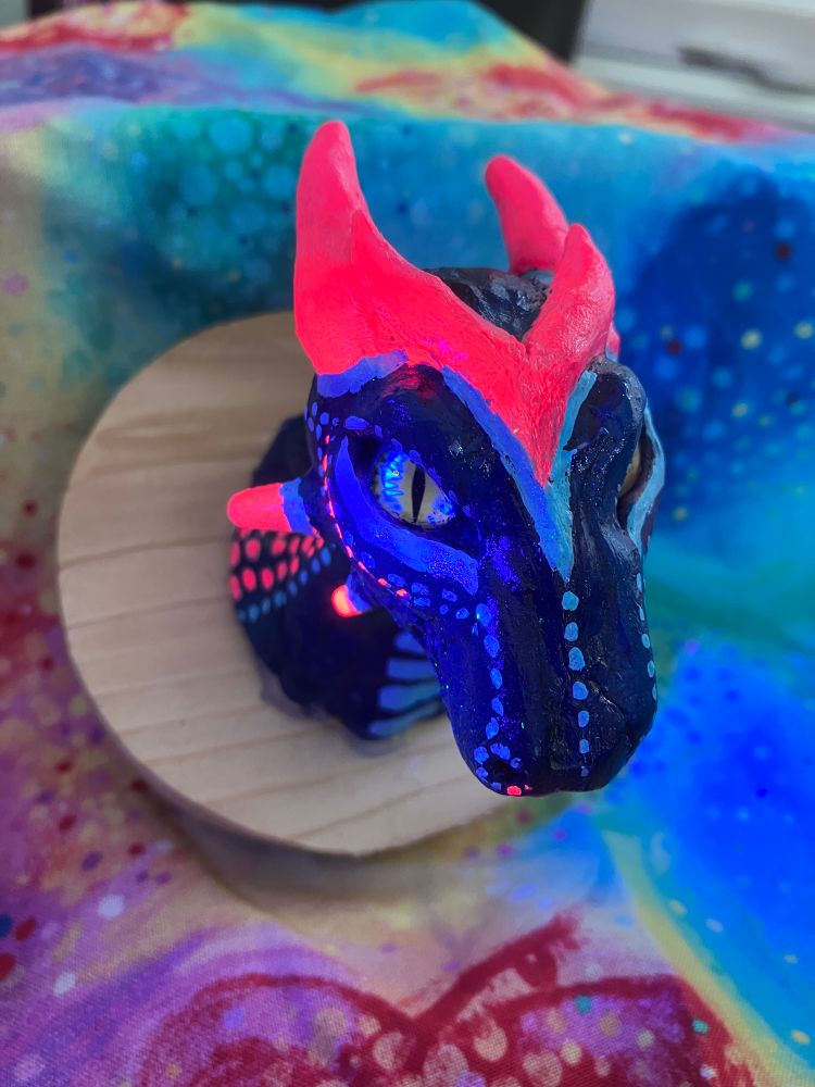 A hand sculpted dragon made of polymer clay, painted with acrylic paint, and mounted on a recycled wood board. Done in a faux taxidermy head mount style. It is under a blacklight in this photo