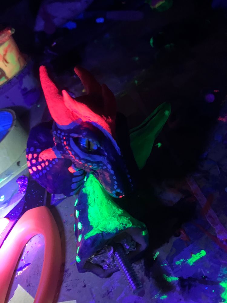A wip picture of a hand sculpted dragon made of polymer clay and painted with acrylic paint. Done in a faux taxidermy head mount style. It is under a blacklight in this photo