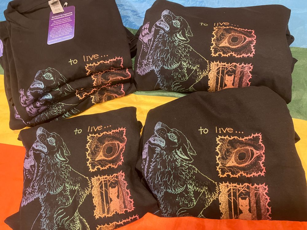 A collection of handprinted (hand screen printed) shirts in rainbow ink featuring a transmasc werewolf
