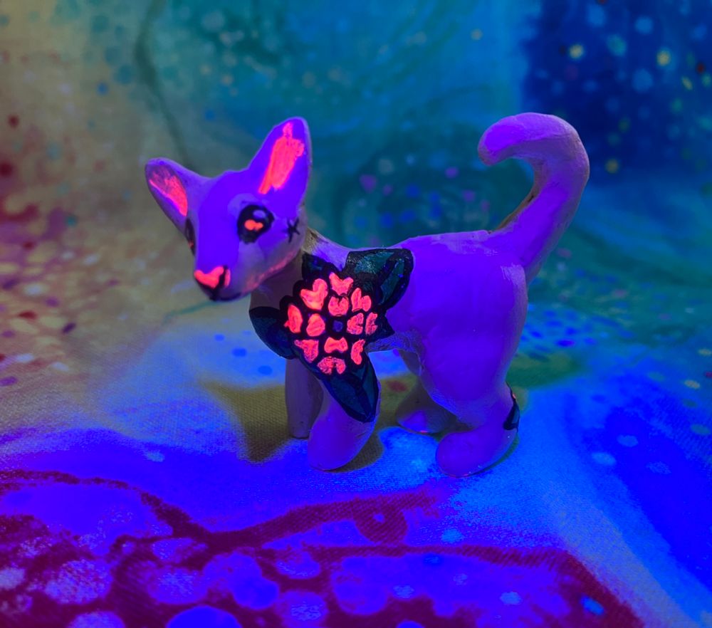 A mini polymer clay sculpture of a sphynx cat with uv reactive floral tattoos painted on with acrylic paint under a black light