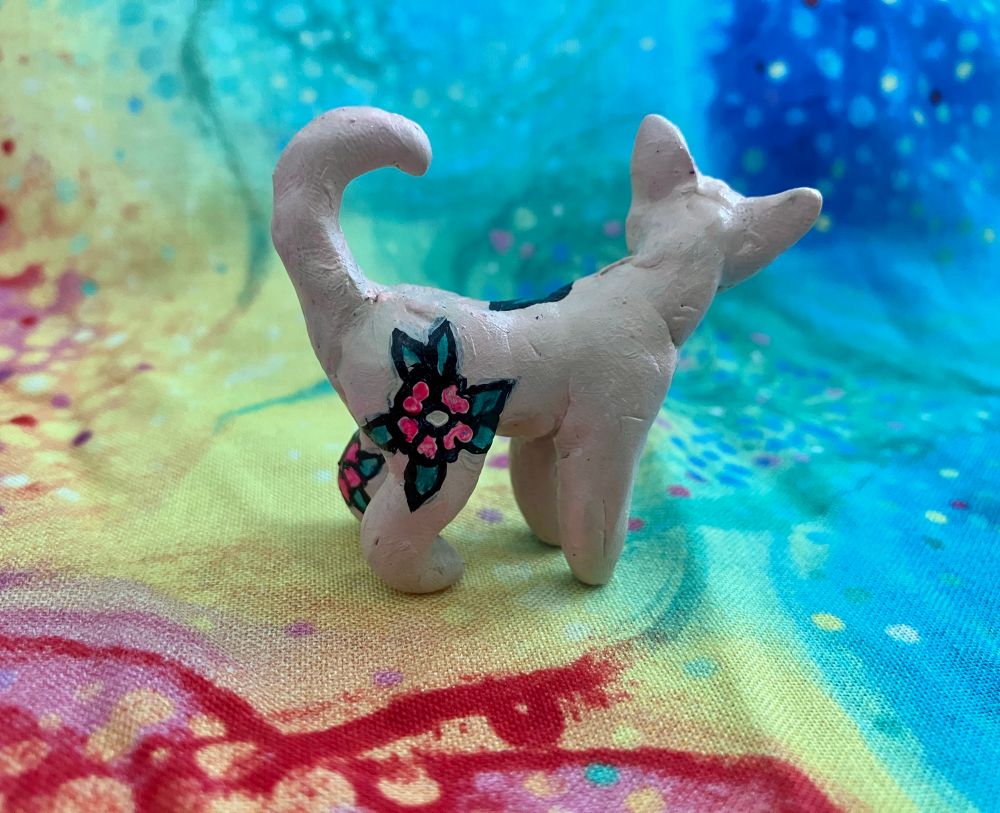 A mini polymer clay sculpture of a sphynx cat with uv reactive floral tattoos painted on with acrylic paint