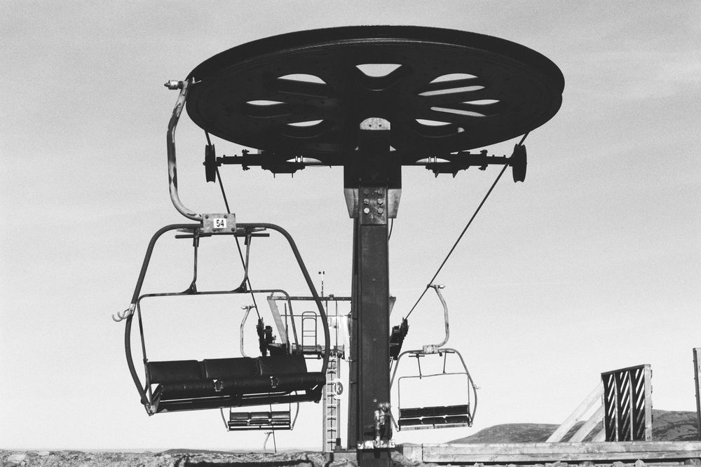 Black and white photograph at the top of a three-abreast chairlift, the top pulley and mast are shown.